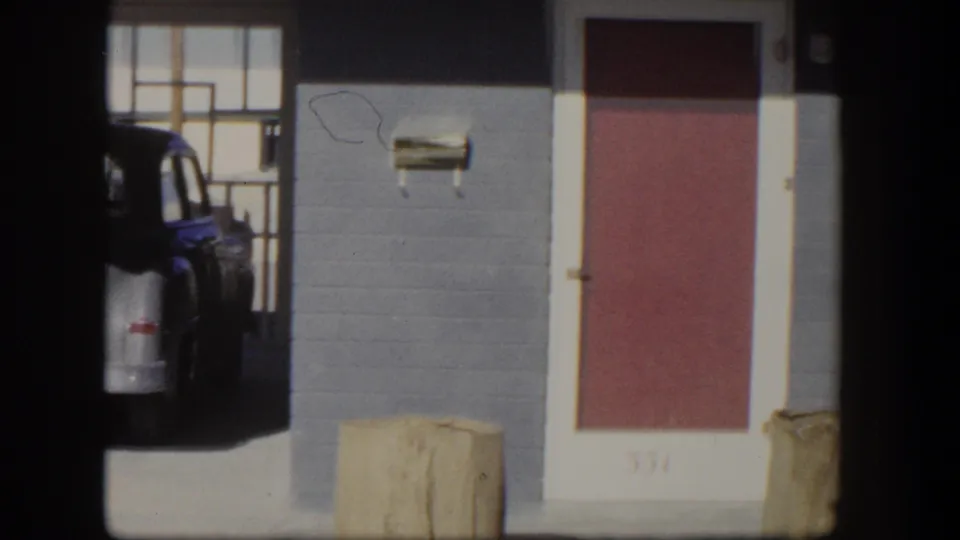 1954 Nevada 8mm frame with a red door, gray residential wall, white trim, tan container, and a partially visible blue car.