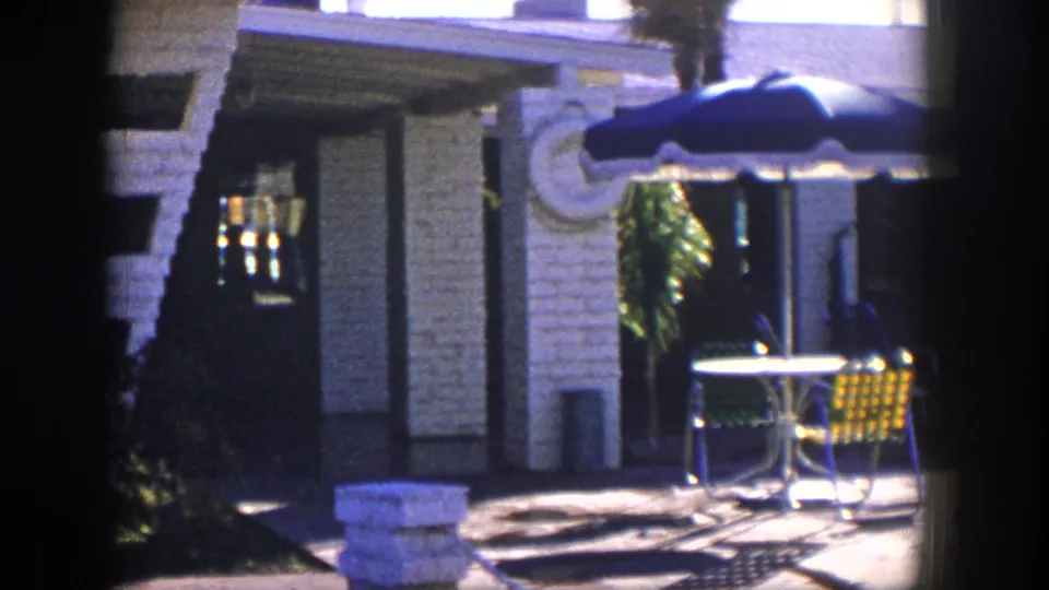1954 Nevada poolside patio area with a blue umbrella, metal seating, round table, stone facade, and bright afternoon light.