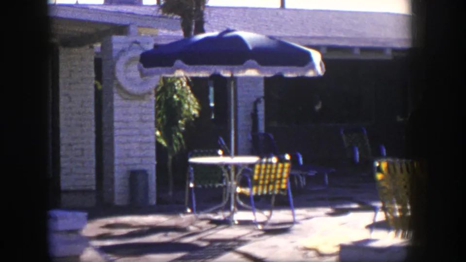1954 Nevada home-movie view of poolside patio furniture under a blue umbrella beside a stone motel facade.