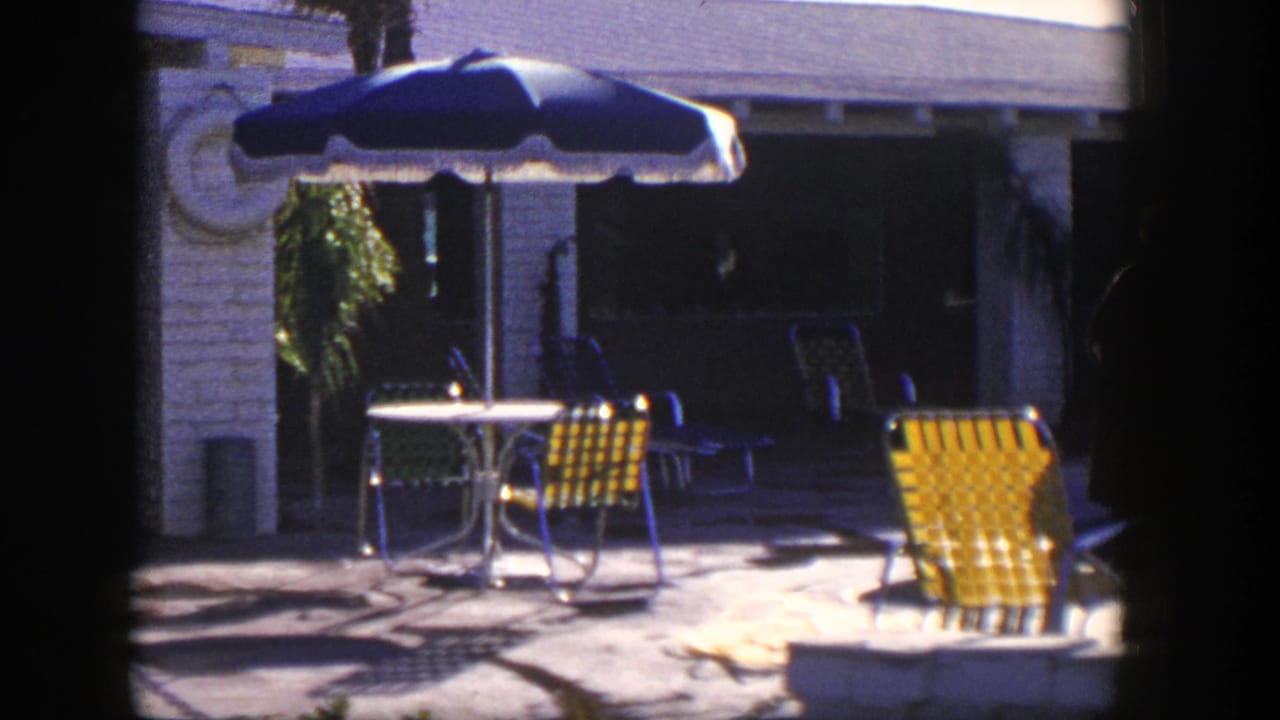 1954 Nevada 8mm frame showing a bright patio with yellow woven chairs, blue umbrella and shaded building frontage.