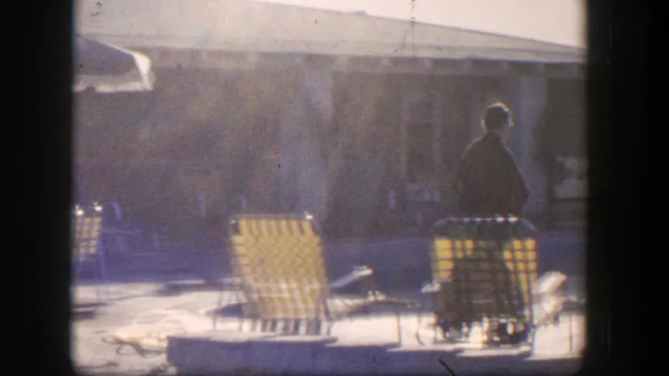 1954 Nevada home-movie frame with a lone adult in a dark coat crossing a patio near yellow chairs and a low building.