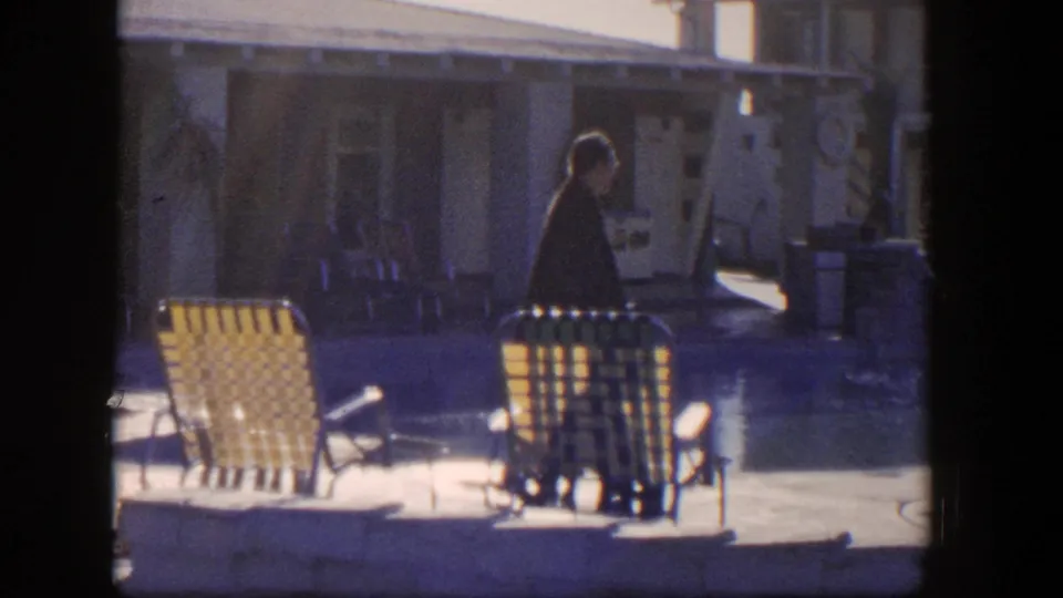 1954 Nevada 8mm frame showing a lone adult near yellow woven chairs, with a shaded building and doorway in the background.