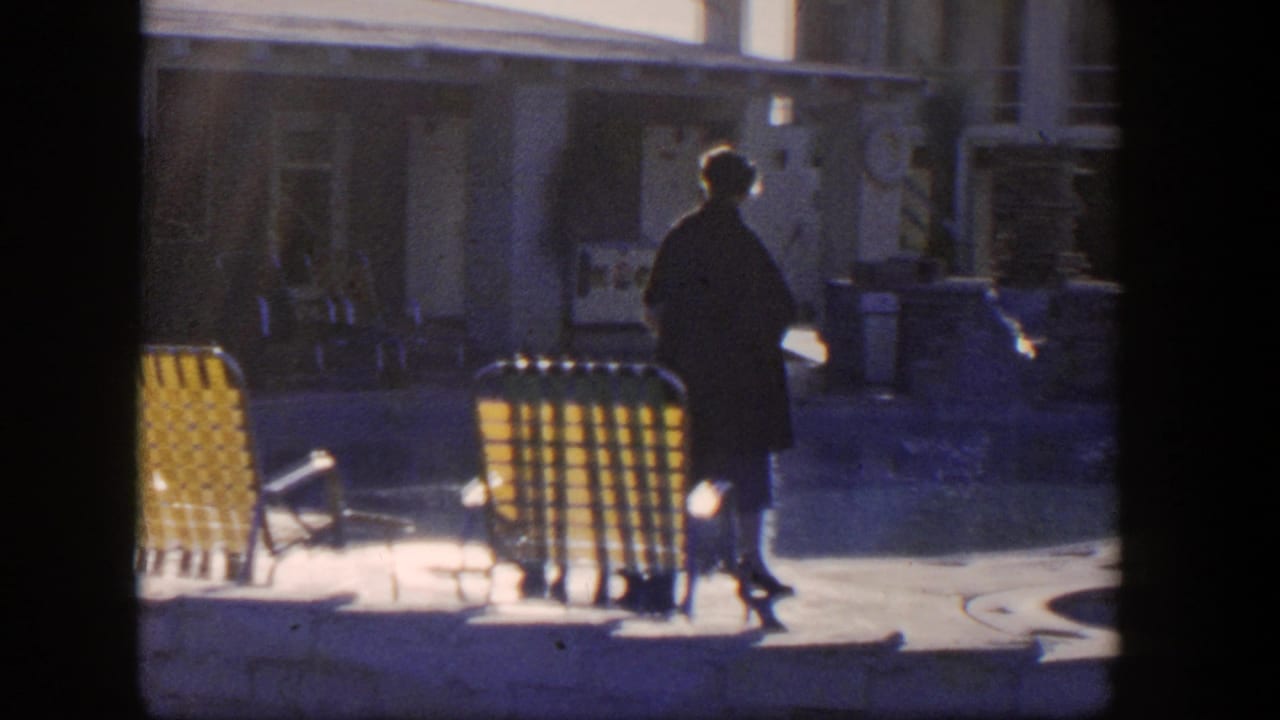 Woman in a dark coat walks beside a motel swimming pool near yellow lounge chairs.