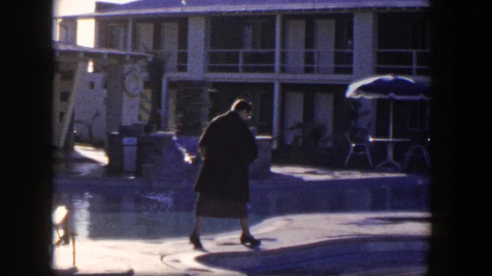 A woman in a dark coat passes the motel swimming pool, with a patio umbrella, railings, and shaded rooms in the background.