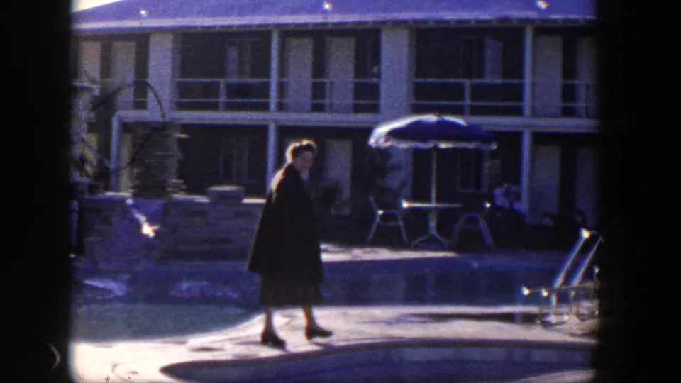 Near the end of the Nevada poolside shot, the woman appears near the pool edge with motel balconies and a patio umbrella behind her.