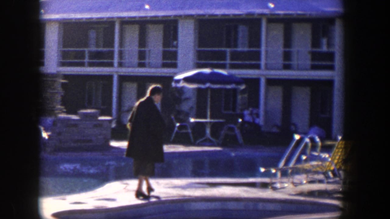 In 1954 Nevada home-movie footage, a woman in a dark tweed-style coat walks near a sunlit motel pool and patio furniture.