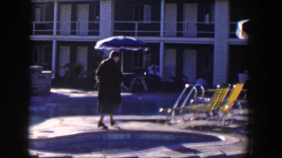 The woman continues along the pool area, framed by yellow lounge chairs, a blue umbrella, and a two-story motel facade.