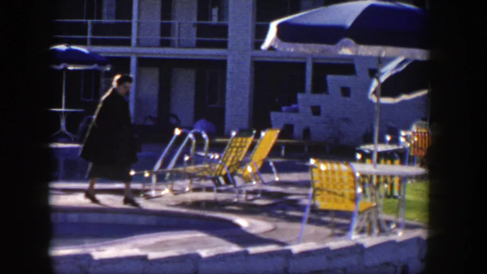 A woman in heels walks beside the pool, passing metal lounge chairs, a pool ladder, patio seating, and motel rooms.