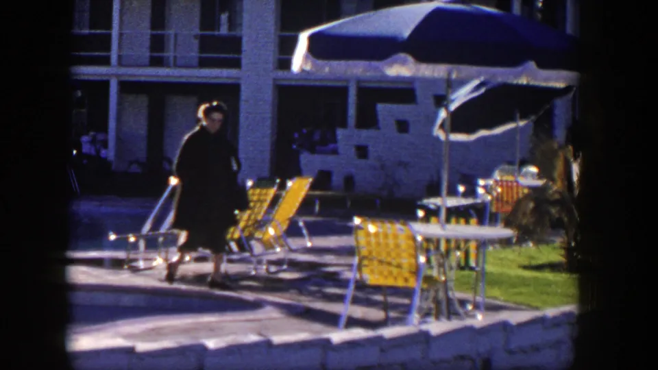 The woman passes the poolside seating area, with yellow chairs, a blue umbrella, grass, and the motel building visible behind her.