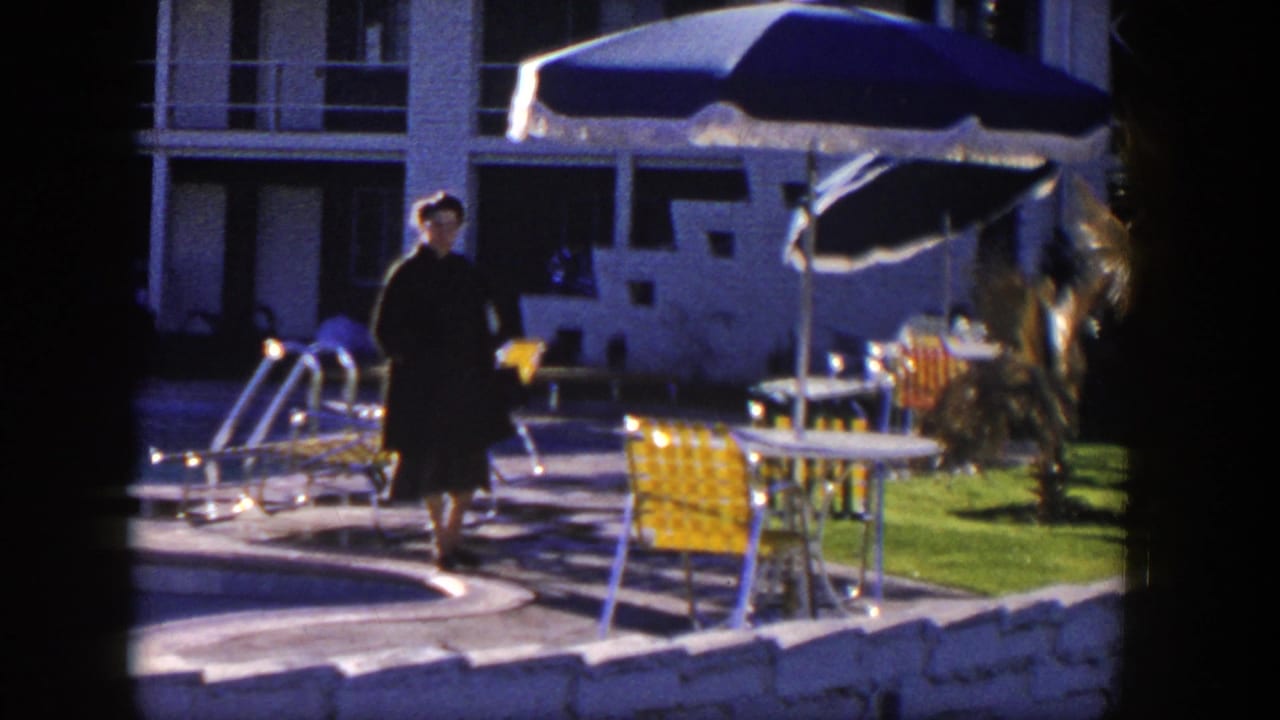In 1954 Nevada home-movie footage, a woman in a dark dress walks beside a pool patio with blue umbrellas and yellow chairs.