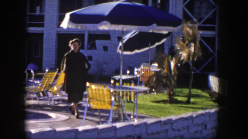 The woman continues along the Nevada poolside walkway, passing yellow chairs, tables, umbrellas, and a modern building facade.