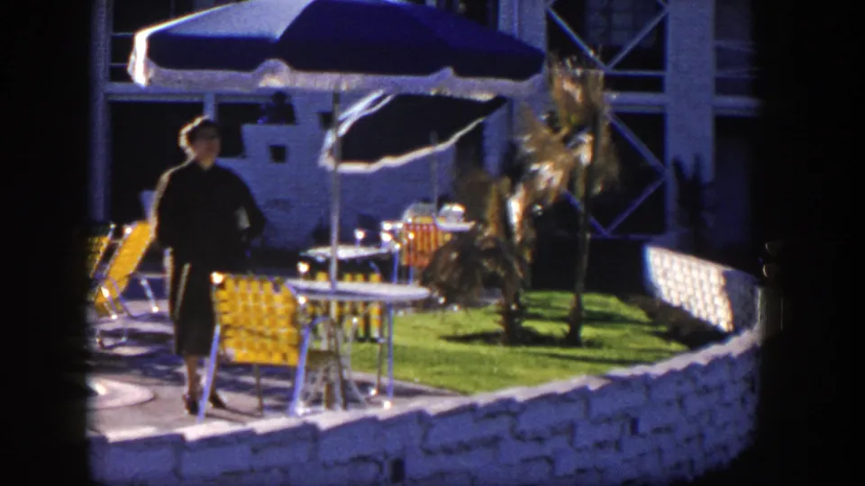 A woman in a dark dress passes near yellow patio chairs, with blue umbrellas, a small lawn, palm plantings, and a stone wall visible.