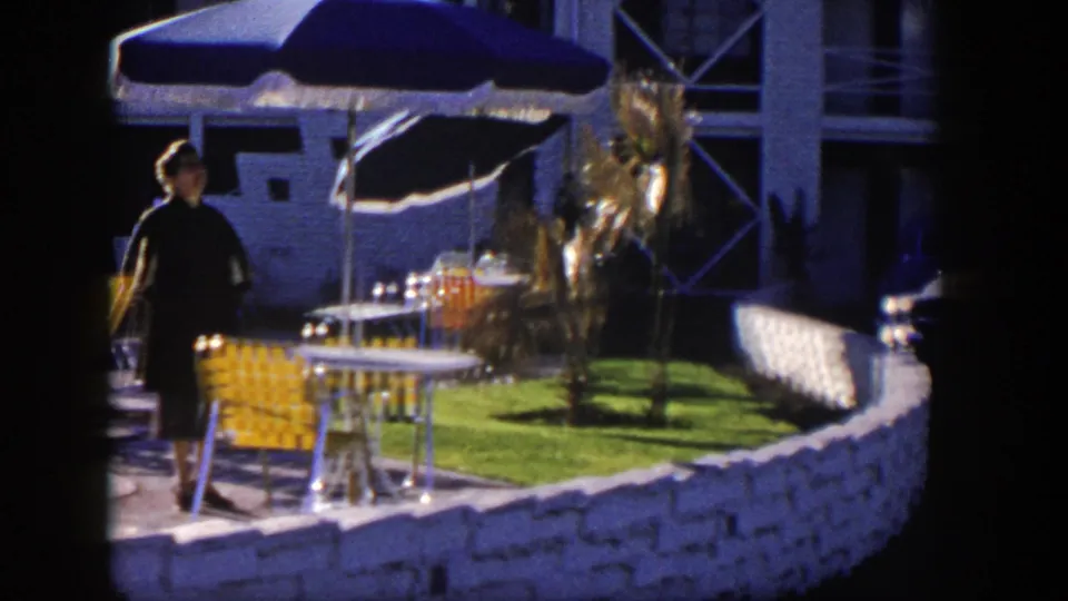 Near the end of the shot, the woman walks closer to camera beside poolside seating, umbrellas, lawn, palm plantings, and a low stone wall.
