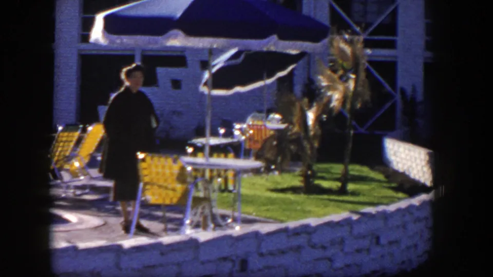 1954 Nevada 8mm frame showing a woman by yellow patio chairs, blue umbrellas, a small lawn, and a white stone fence.