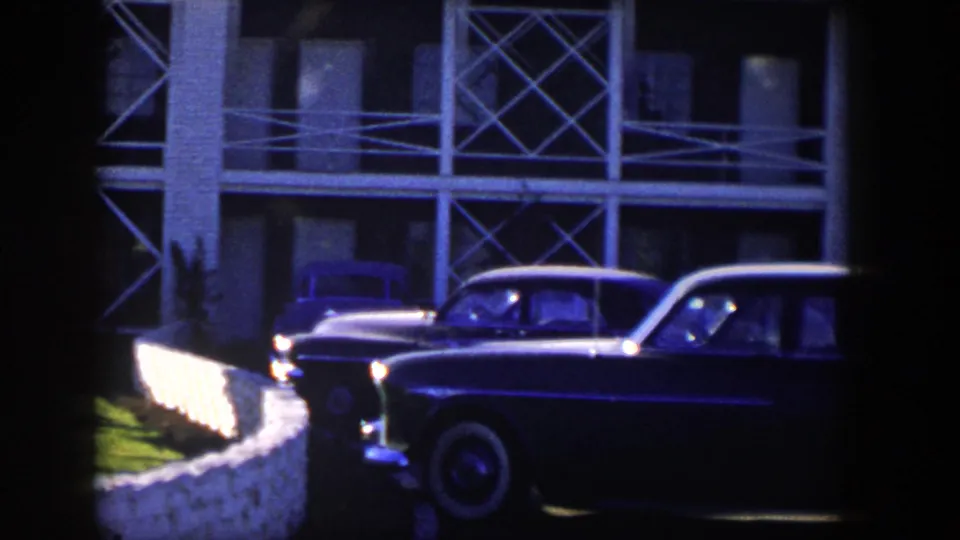 1954 Nevada home-movie frame showing a dark mid-century car parked along a white stone fence near a multi-window building.