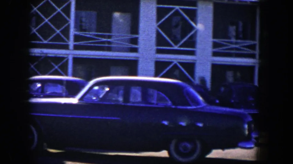 1954 Nevada frame with a dark parked sedan crossing the foreground before a building with geometric balcony railings.