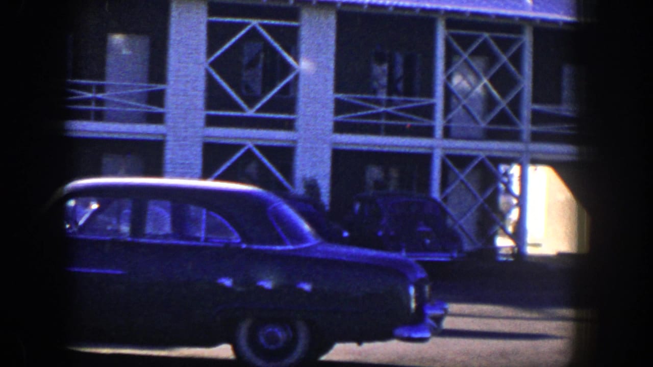 Parked sedan in front of a multi-story building with geometric balcony rails.