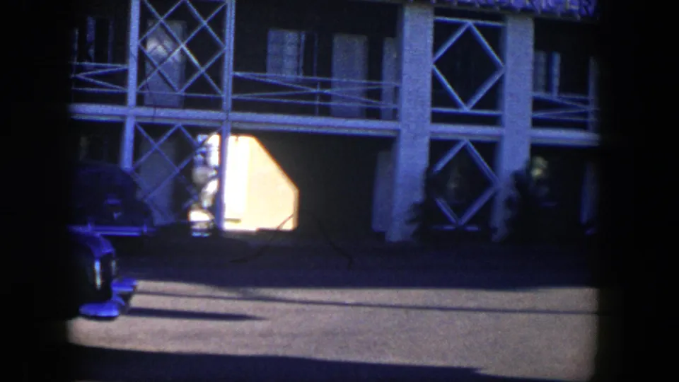 1954 Nevada 8mm frame of a modernist-looking building facade, bright doorway, steps, and part of a parked car.