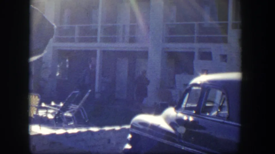 1954 Nevada 8mm frame showing lounge chairs, a parked car, and a distant person near the building exterior.