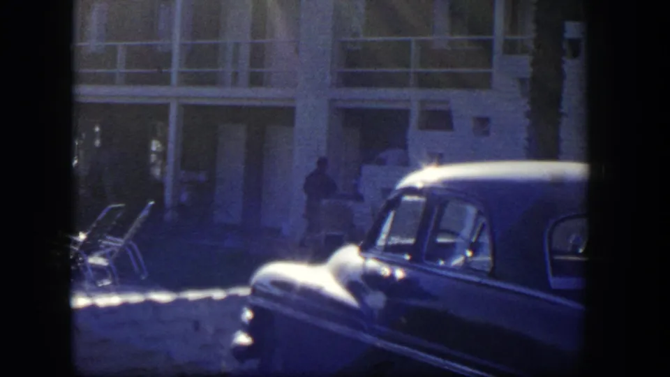 1954 Nevada 8mm frame with a parked car in the foreground, lounge chairs, and a distant person by the building.