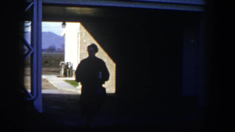 1954 Nevada 8mm frame of a silhouetted person near a shaded doorway, with sunlit ground and distant hills visible beyond.