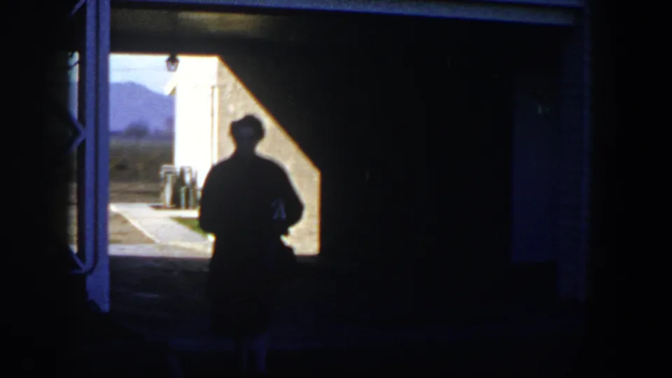 In 1954 Nevada home-movie footage, a woman in silhouette walks from a shaded garage opening toward a sunlit yard with mountains beyond.