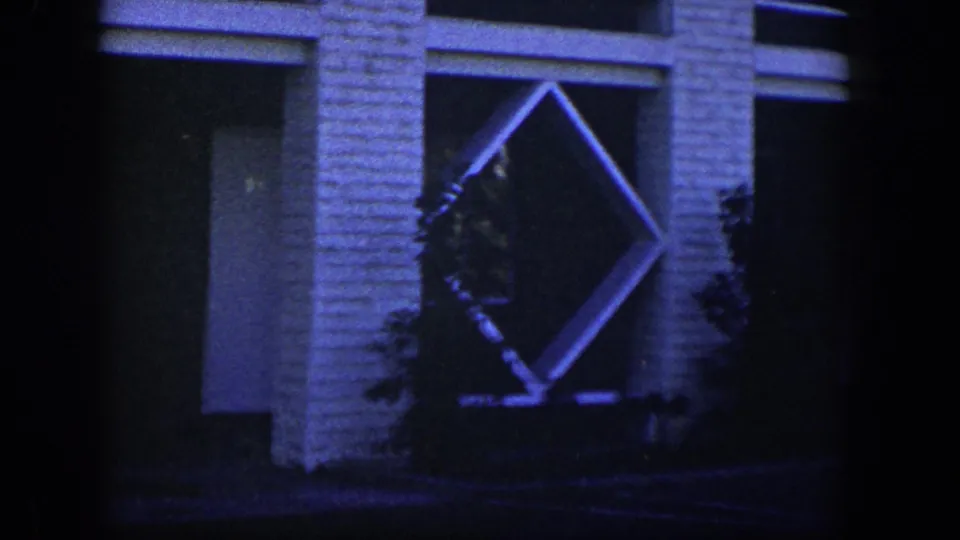 1954 Nevada 8mm frame showing a white brick building facade with a diamond lattice feature, dark window areas, and shrubs in blue evening light.