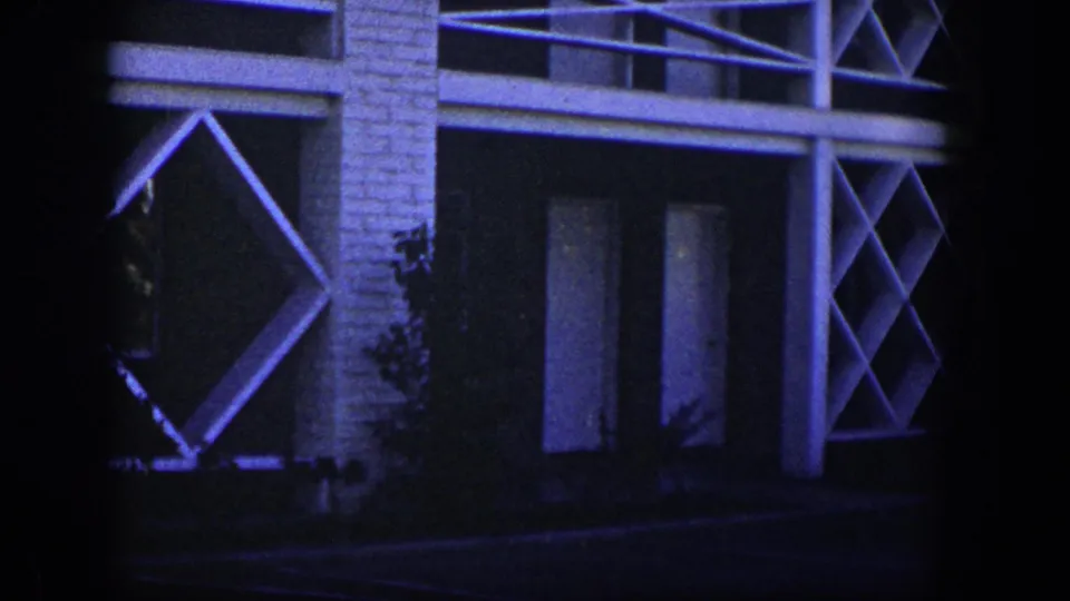 1954 Nevada 8mm frame showing more of the facade, including a diamond lattice panel, vertical supports, dark windows, shrubs, and a paved foreground edge.