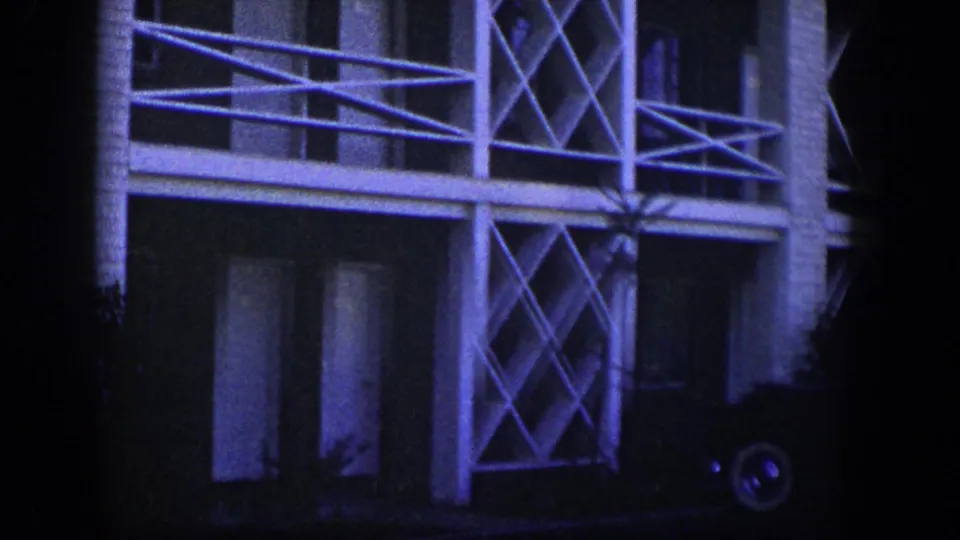 1954 Nevada frame showing a white brick facade with crossed lattice panels, horizontal rails, dark windows, shrubs, and part of a vehicle wheel at the edge.