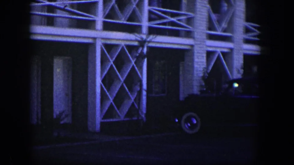 1954 Nevada 8mm frame showing a dark classic sedan beside a brick building with white cross-lattice balcony details.