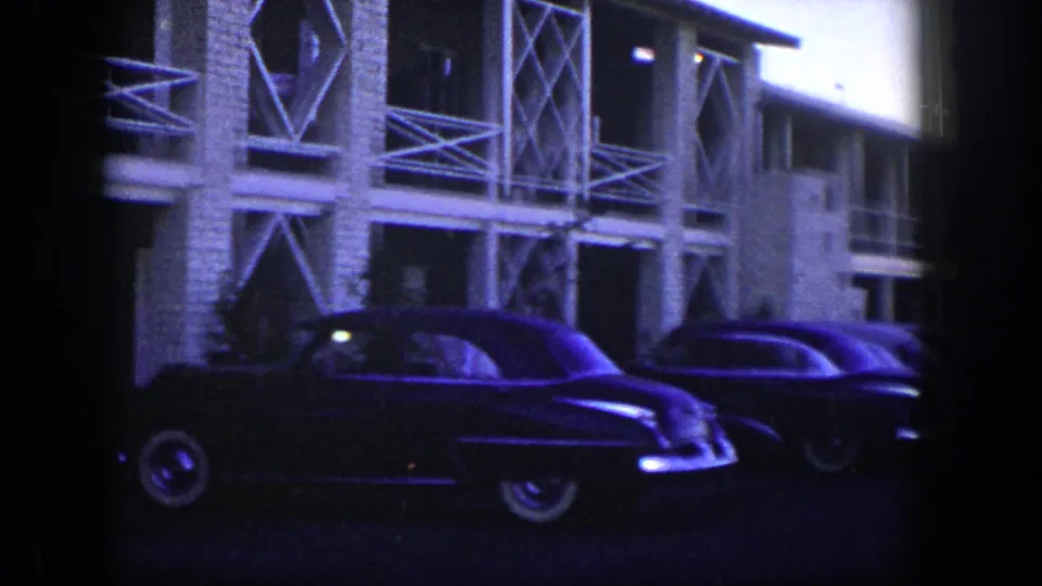 1954 Nevada home-movie frame with two parked dark sedans below a two-story building with white lattice rails.