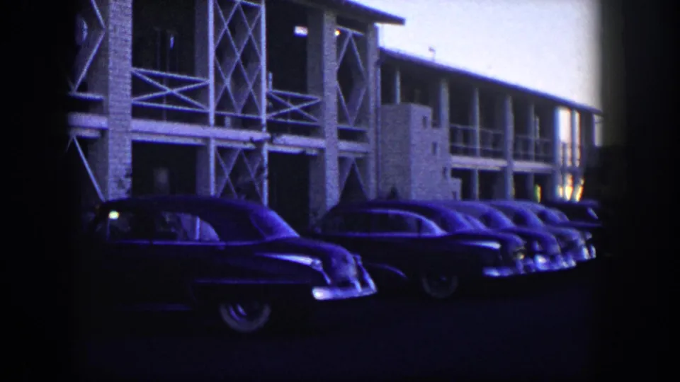 1954 Nevada 8mm frame showing parked mid-century cars in shadow beside a motel with cross-braced supports.