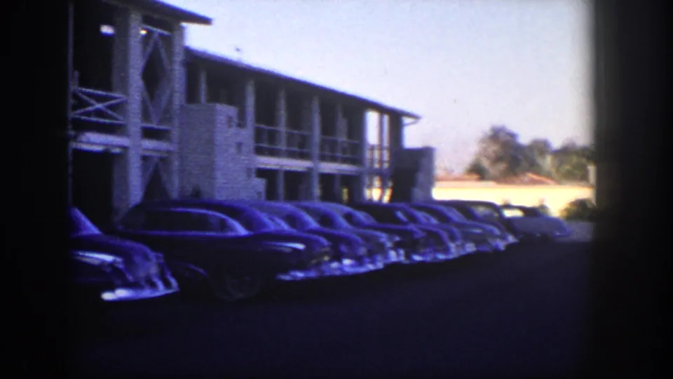 1954 Nevada roadside scene with rounded sedans parked along a mid-century motel, seen through blue-purple film shadows.