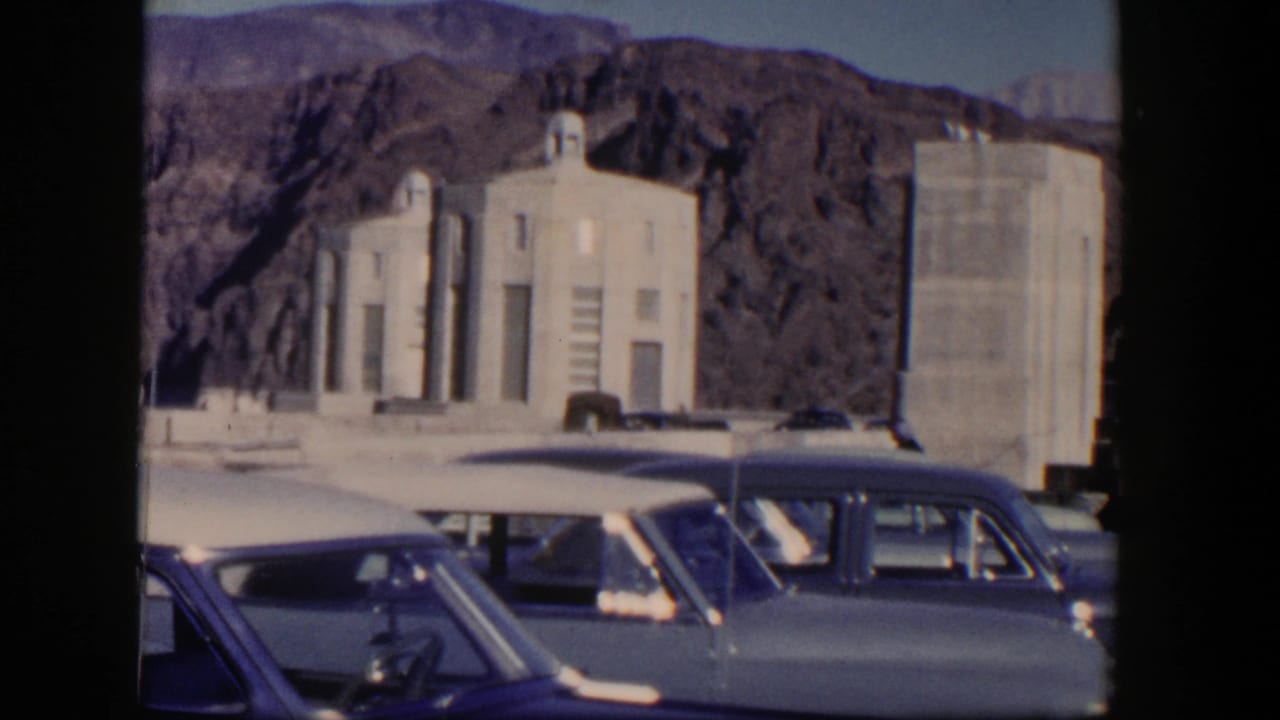 1954 Nevada 8mm frame showing parked mid-century cars, white stone structures, and rocky desert hills in bright sun.