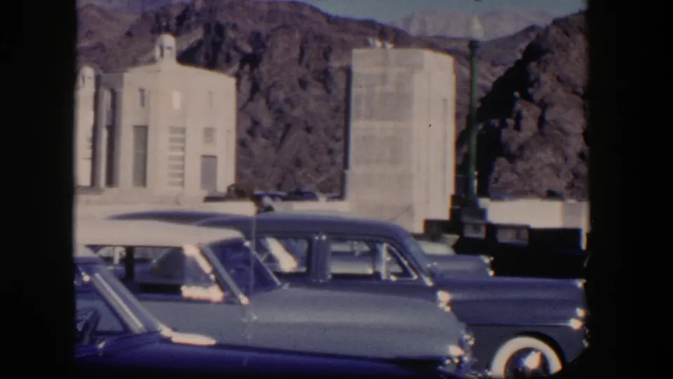 1954 Nevada frame with mid-century parked cars, pale architectural forms, and rugged mountain backdrop.