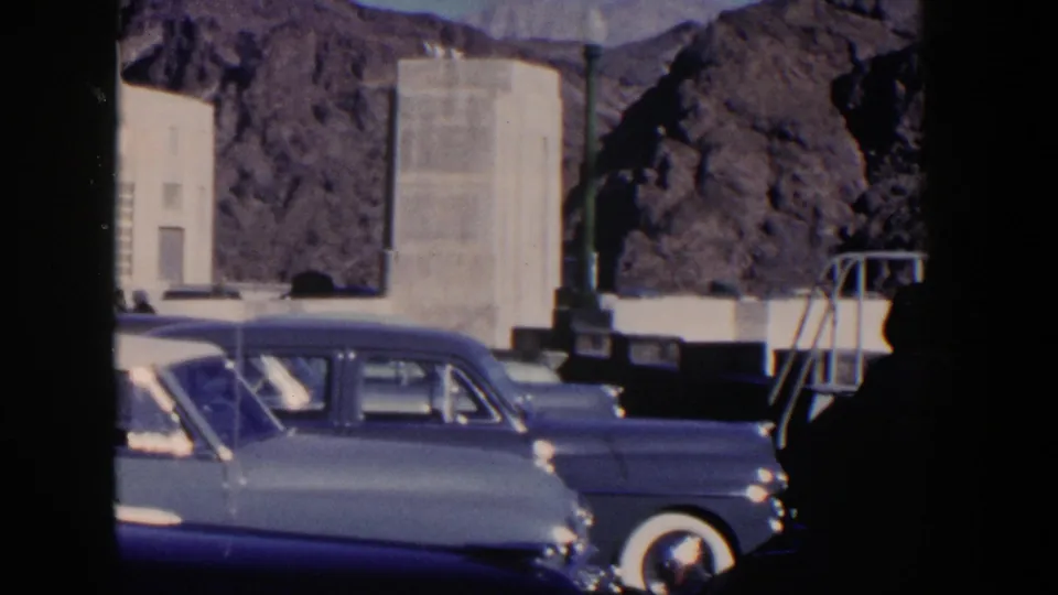 1954 Nevada home-movie frame showing parked sedans beside pale stone architecture with rugged desert rock behind.