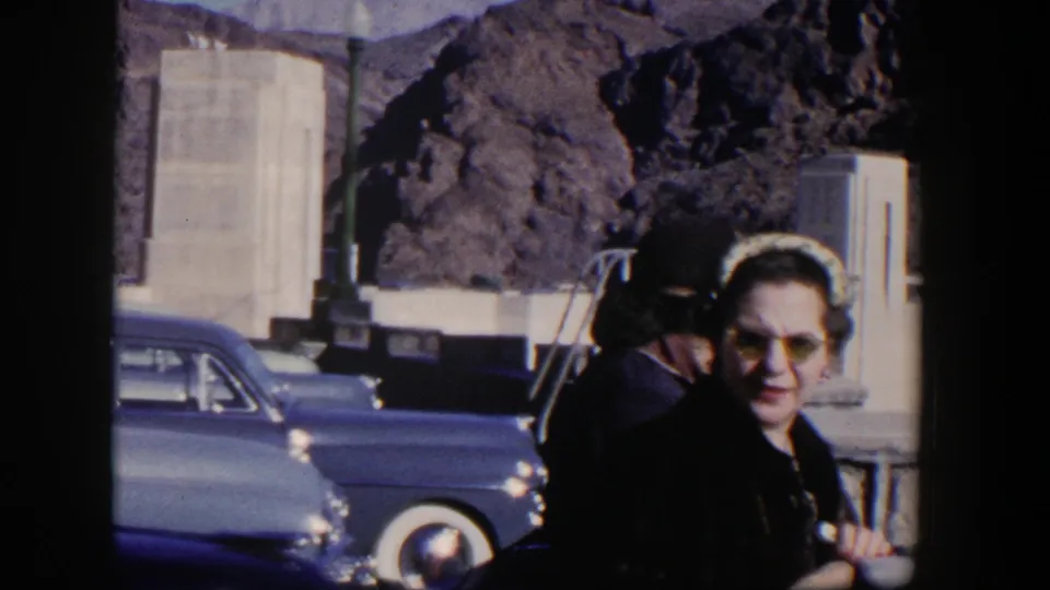 1954 Nevada frame with a woman entering foreground beside parked cars, pale stone structures, and desert mountains.