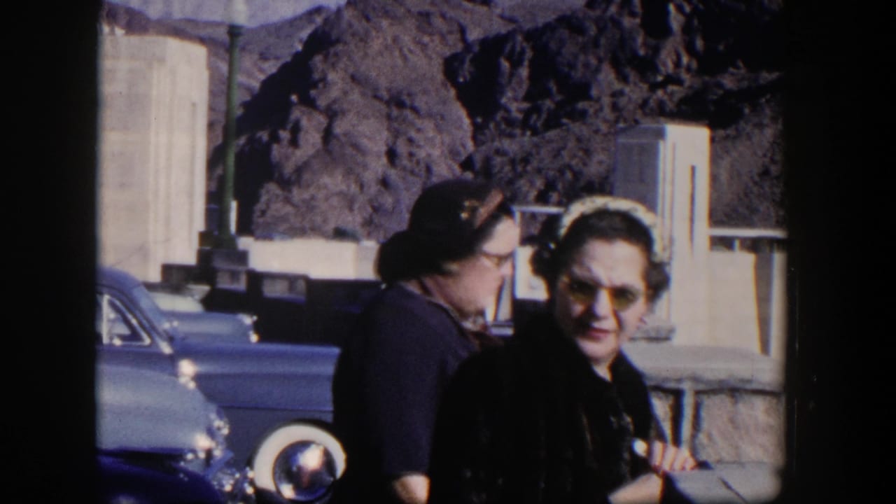 Two women in sunglasses stand near a blue car with concrete dam structures and dark rocks behind them.