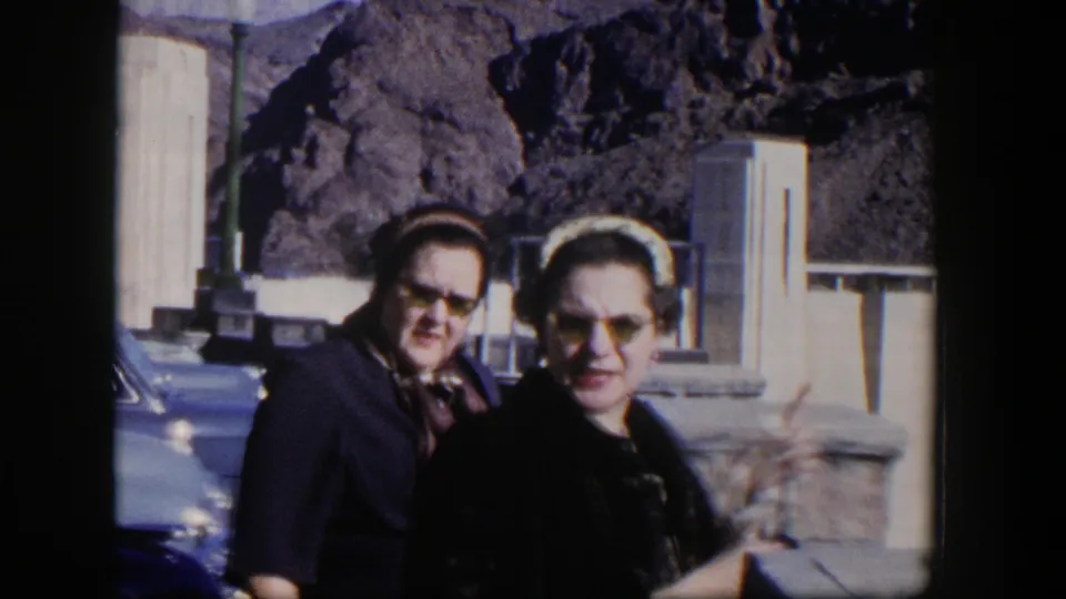 Two women in mid-century attire look toward the camera beside a blue car, with concrete dam features and rocky Nevada terrain behind them.