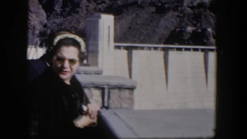 A woman in sunglasses stands partly cropped in a 1954 Nevada home-movie view, with dam concrete and desert rock in the background.