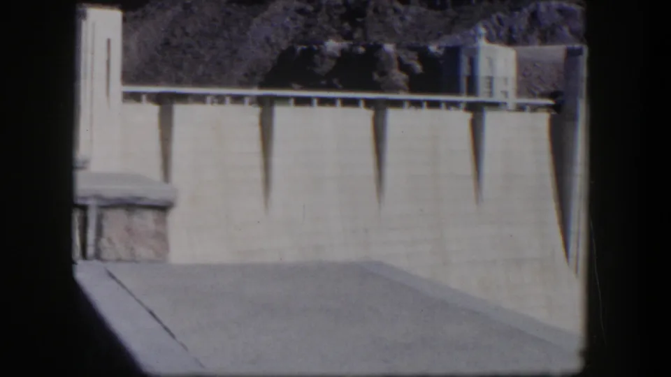 The camera drifts away from the figures to show concrete dam structures, pale railings, desert rock, and a sunlit roadway in Nevada.