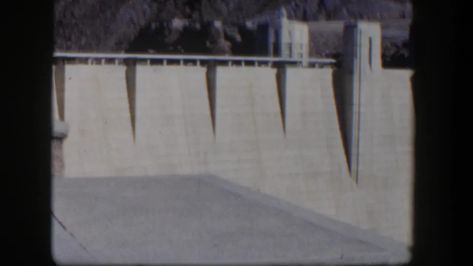 1954 Nevada 8mm frame showing a close view of Hoover Dam's pale concrete face, shadowed vertical forms, and dark contact-sheet border.