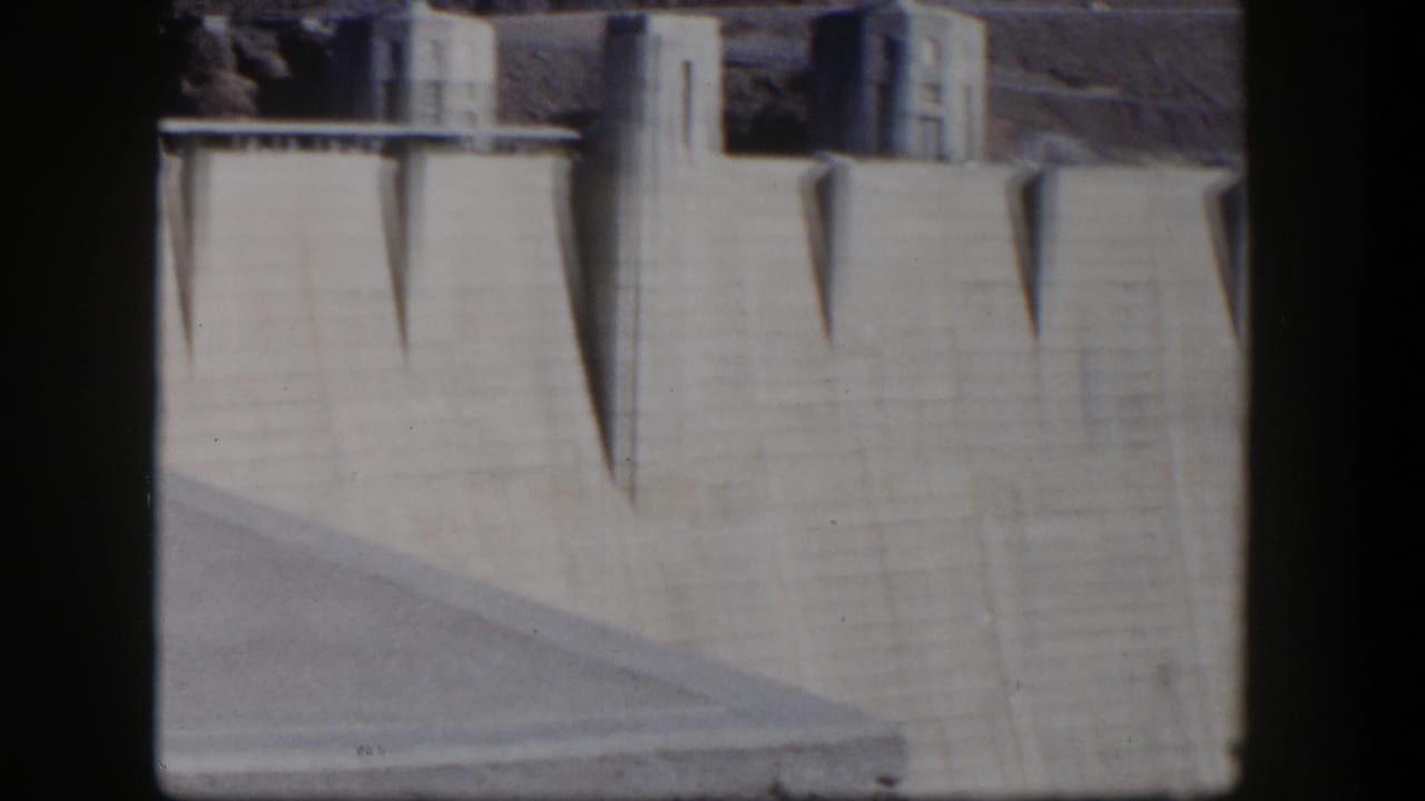 Concrete dam face with upper structures, sharp shadows, and dark frame edges.
