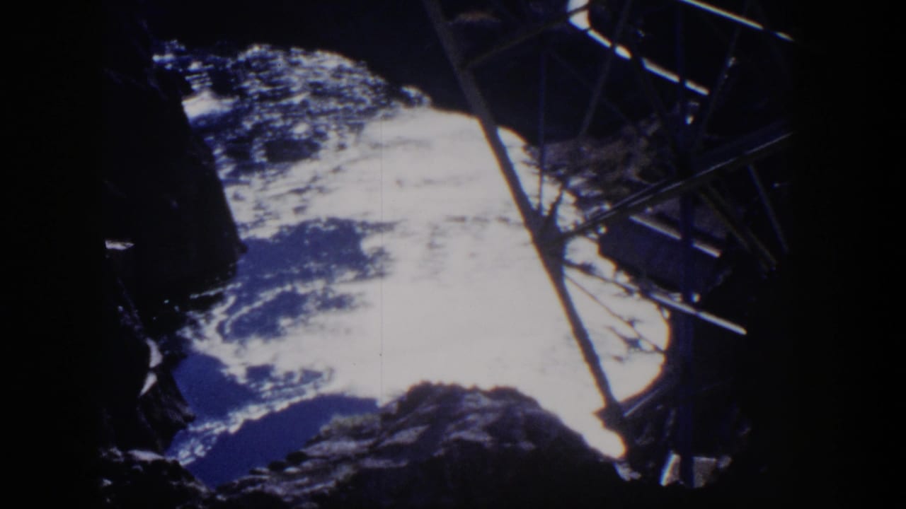 1954 Nevada 8mm frame showing jagged dark rock, bright clouded sky, and a metal support intruding from the right edge.