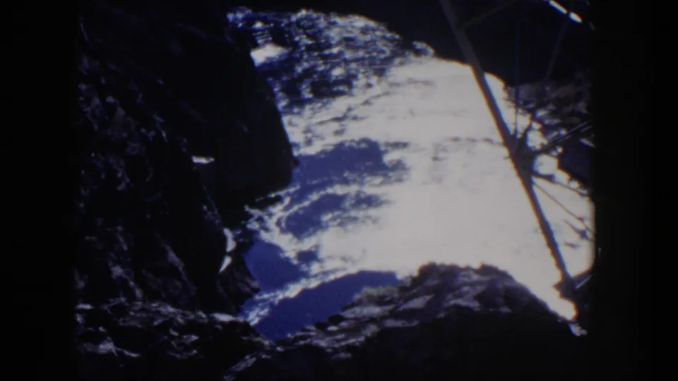 1954 Nevada 8mm view across dark foreground rock toward pale clouds and distant blue terrain, crossed by a diagonal metal element.
