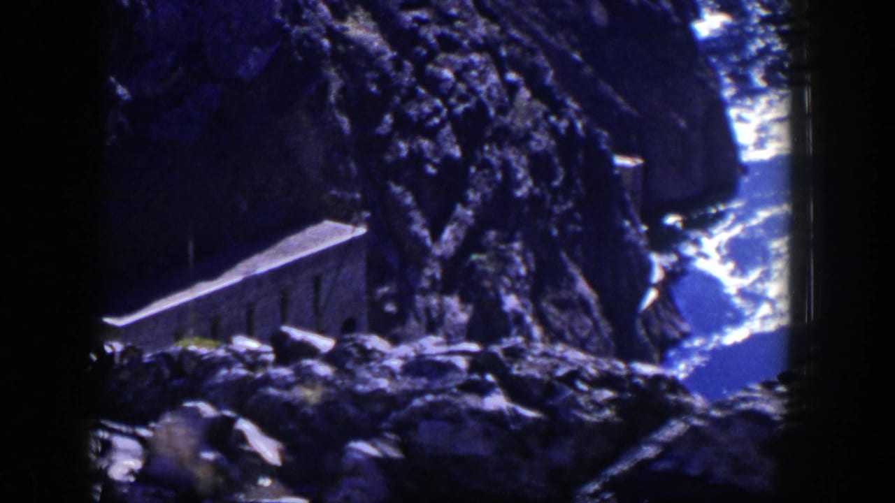 Small stone building set among steep dark rocks near bright water.