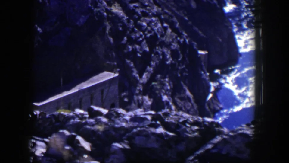 1954 Nevada home-movie frame with a small stone building tucked into a rocky cliffside above bright water.