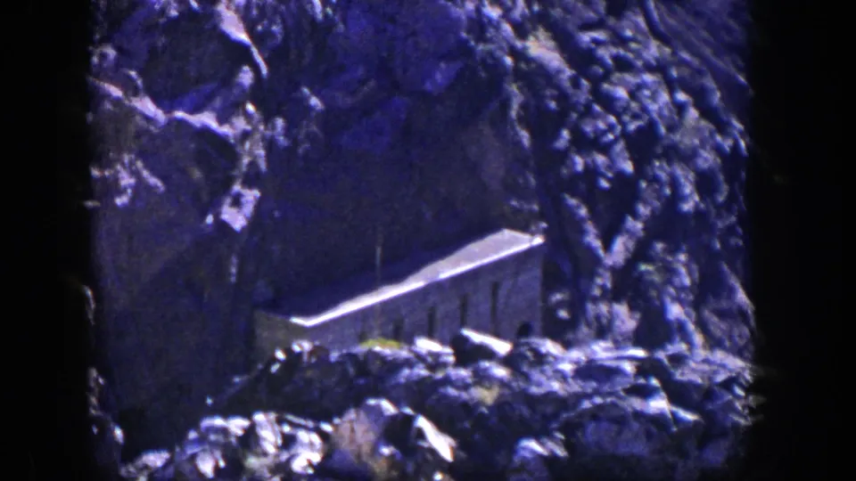 1954 Nevada 8mm frame showing rough cliff texture and a small cliffside building partly obscured by shadow.