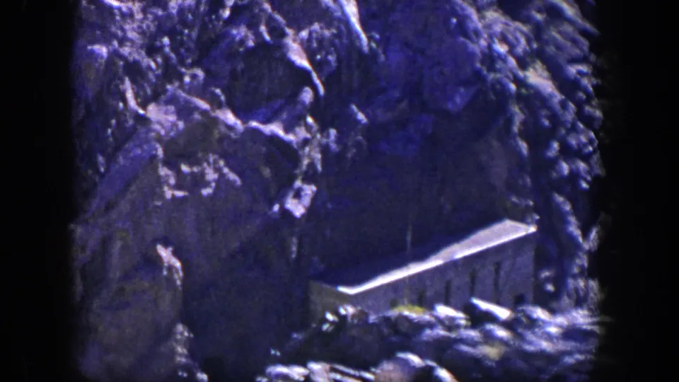 1954 Nevada home-movie frame showing a small stone building beneath a steep blue-purple cliff face.