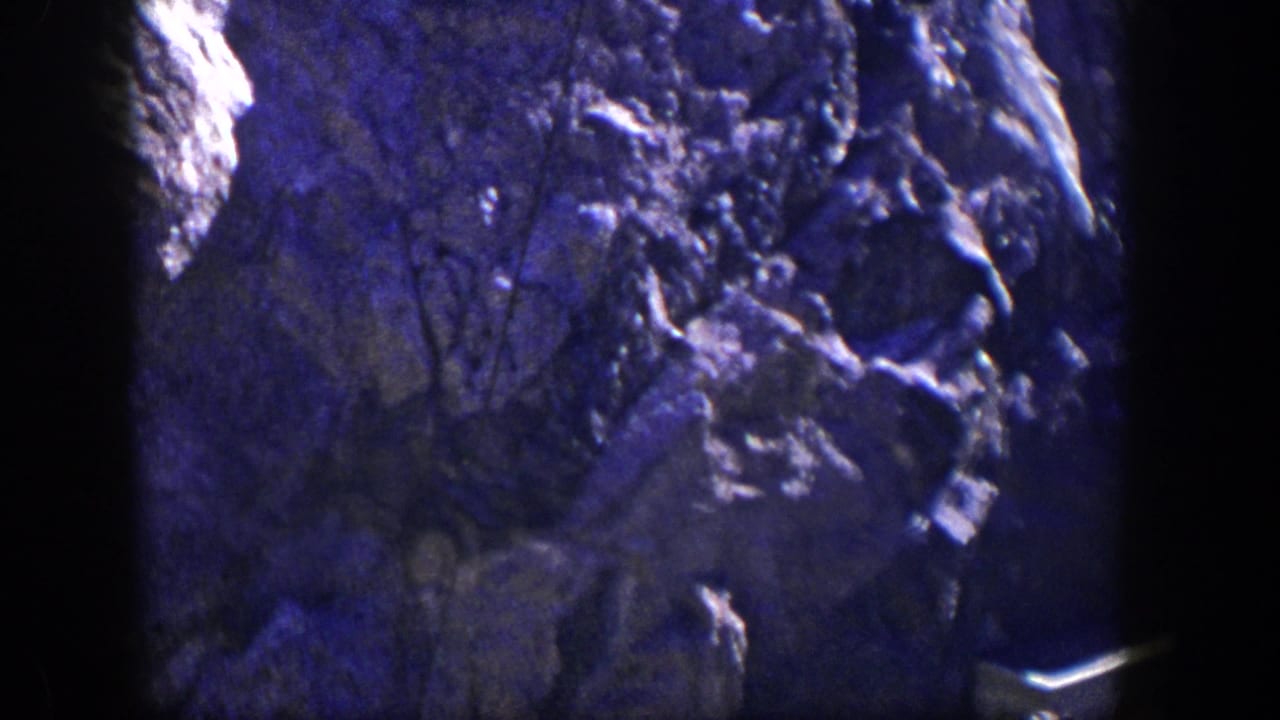 Rock wall with a dark central border, sparse brush silhouettes, and a pale structure at lower right.